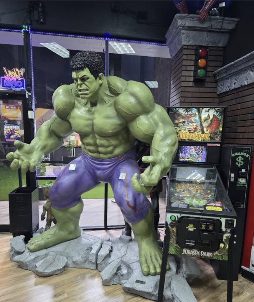 Inside the Gods & Monsters comic and gaming store in Orlando, a large, detailed statue of Marvel’s Hulk stands mid-roar on a cracked stone base. The green superhero wears torn purple pants and is surrounded by arcade machines, including a Jurassic Park game, under bright indoor lighting.