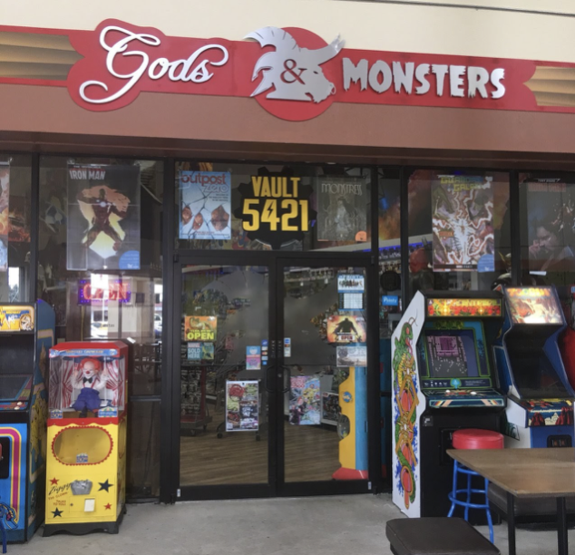 The storefront of Gods & Monsters, an Orlando comic and gaming shop, featuring a red sign with white lettering above glass doors. Arcade cabinets and colorful machines line the entrance under a covered outdoor walkway.