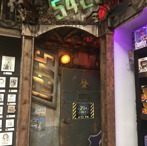 A themed interior room at Gods & Monsters designed like a distressed metal vault, marked with the number 5421. The industrial door features warning symbols and dramatic lighting, with comic art displayed on the surrounding walls.