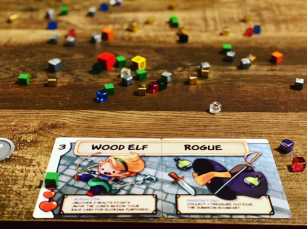 A close-up of a Dungeon Drop player card showing the characters ‘Wood Elf’ and ‘Rogue,’ placed on a wooden table with numerous colorful cubes scattered in the background.