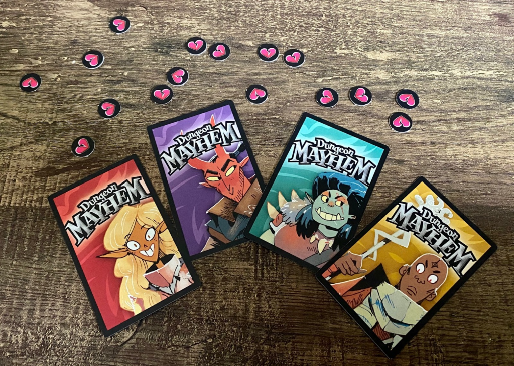 Four Dungeon Mayhem character cards fanned out on a wooden table, with small heart-shaped damage tokens scattered above them. Each card features colorful cartoon-style fantasy characters.