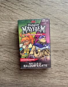 The Dungeon Mayhem: Battle for Baldur’s Gate expansion pack box resting on a wooden surface, featuring illustrated Dungeons & Dragons characters and the label ‘Age 8+’ on the front.