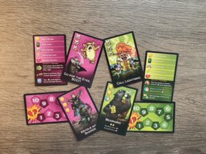 A spread of Dungeon Mayhem gameplay cards on a wooden table, including action cards, character abilities, and numbered damage trackers in pink and green color schemes.