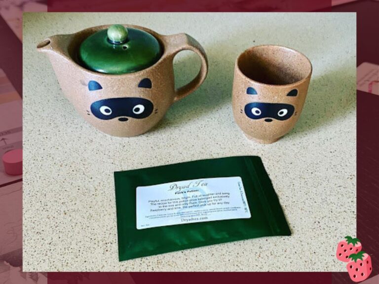 A ceramic teapot and matching cup, featuring tanuki faces are pictured with a green pouch labeled Dryad Tea.