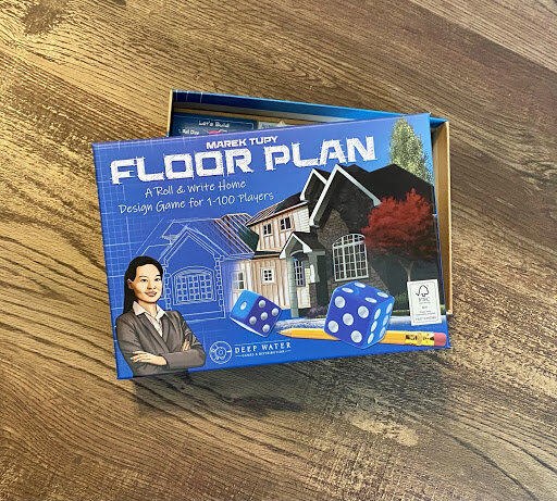 The Floor Plan board game box by Deep Water Games, showing a blueprint-style house illustration, dice, and the subtitle ‘A Roll & Write Home Design Game for 1–100 Players,’ placed on a wooden surface.