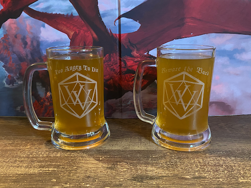Two glass tankard-style mugs filled with a golden-colored beverage, each etched with a tabletop roleplaying dice design and fantasy-themed phrases, set on a wooden surface with a dragon illustration in the background.