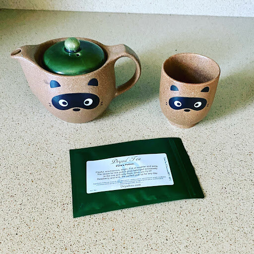A whimsical ceramic teapot and matching cup decorated with a raccoon face sit on a countertop beside a small green packet labeled ‘Dryad Tea,’ showcasing the brand’s playful fantasy aesthetic.