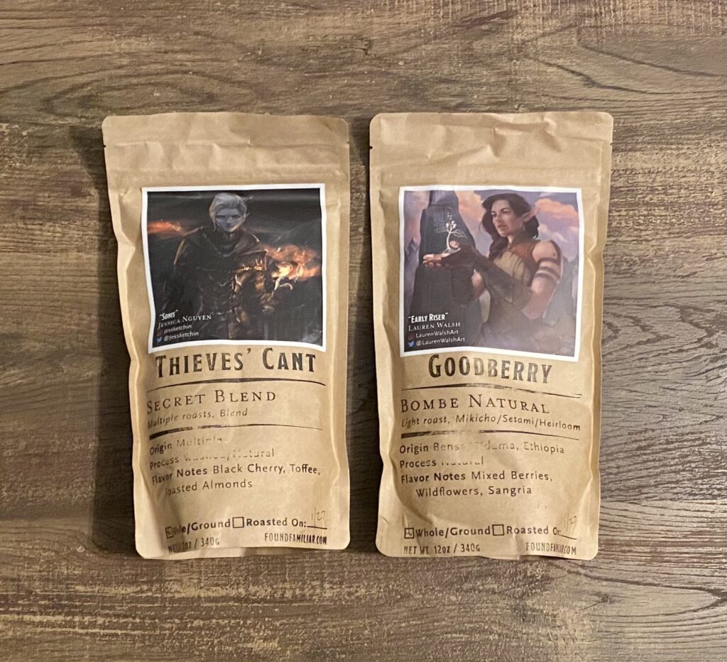 Two bags of Found Familiar Coffee placed side by side on a wooden surface, labeled ‘Thieves’ Cant’ and ‘Goodberry.’ Each bag features fantasy-style character artwork and detailed roast and flavor information on kraft paper packaging.