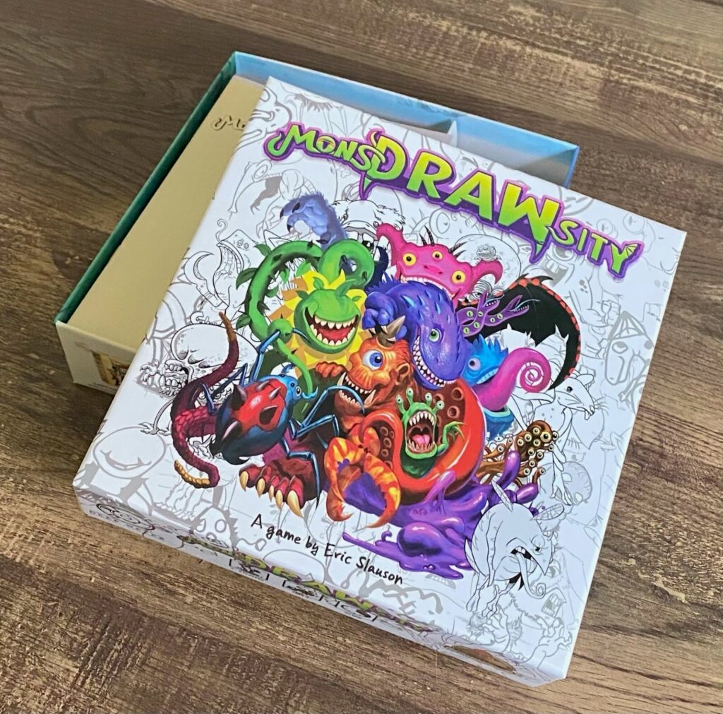 The Mondrawsity board game box partially open on a wooden table, showing colorful cartoon monster artwork on the cover.