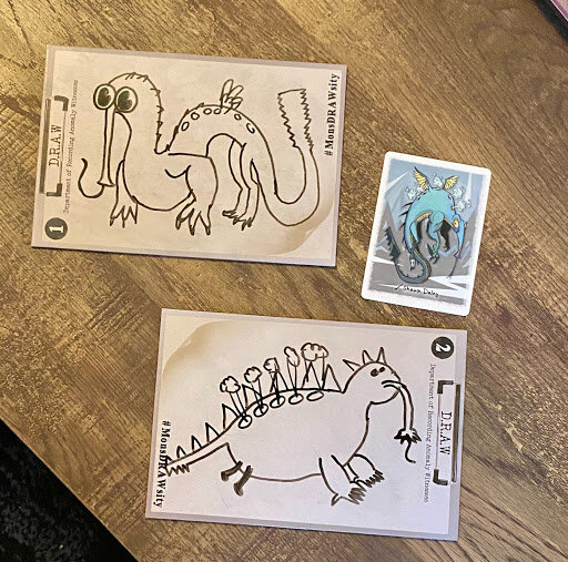 Two Mondrawsity drawing prompt cards with humorous monster sketches alongside a small reference monster card, arranged on a wooden table.