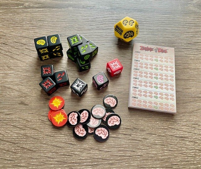 Zombie Dice game components laid out on a wooden table, including custom dice, brain and blast tokens, and a score pad used for tracking player points during gameplay.