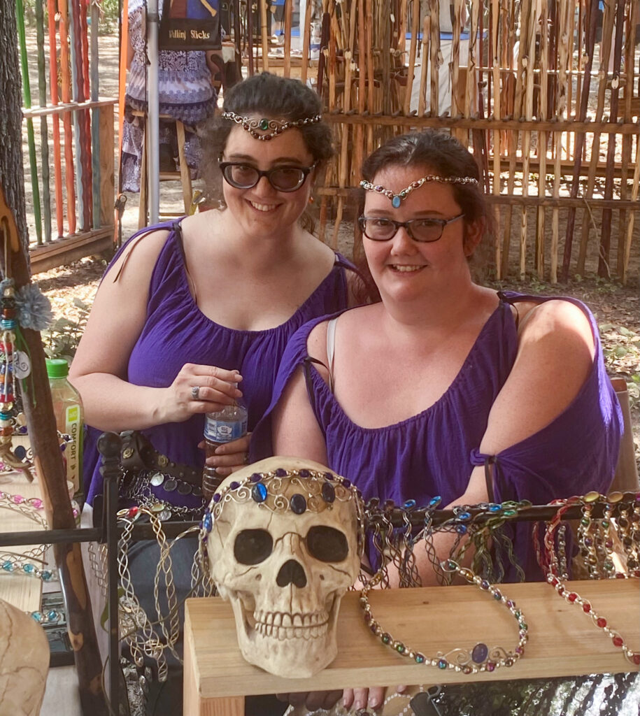 Two women in purple period dresses smile, sporting handcrafted wire circlets that they sell at the Bay Area Renn festival. 