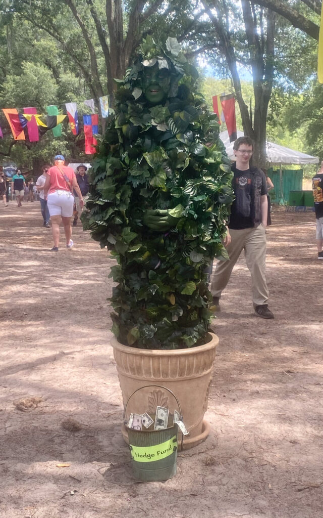 An individual dressed as a bush poses for camera with a tip bucket in the foreground. 