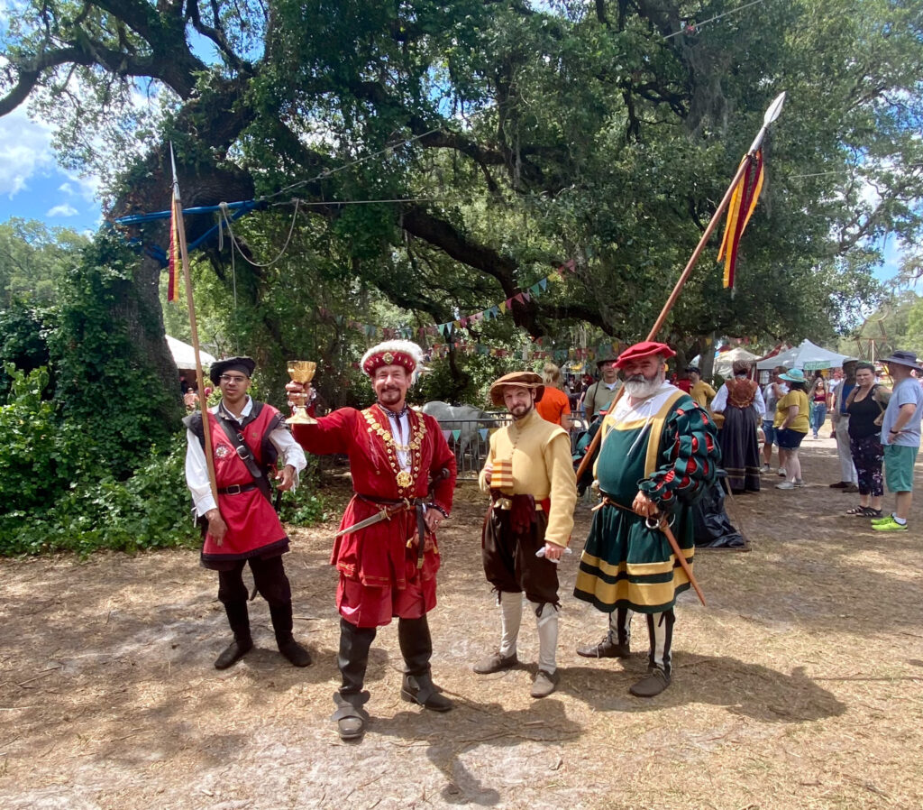 A group of festival actors pose dramatically, with a nobleman dressed in red holding a gold goblet up in toast.
