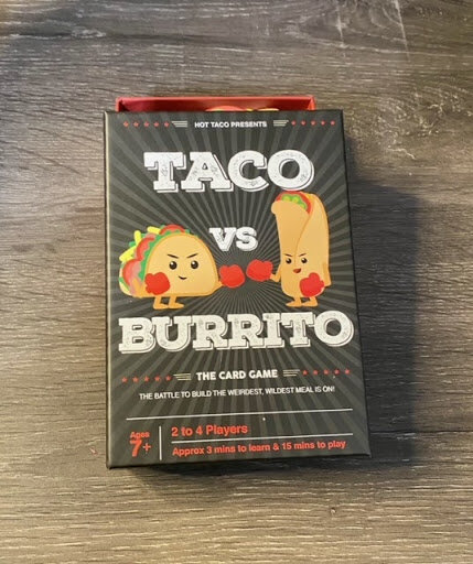 Taco vs Burrito card game box on a wooden table, featuring cartoon taco and burrito characters facing off in a playful boxing match design.