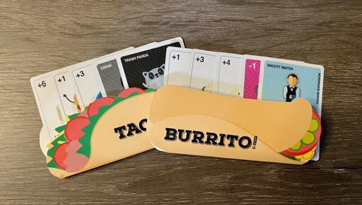 Taco and Burrito player cards displayed in front of a fan of ingredient and action cards, showing the colorful artwork and playful theme of the Taco vs Burrito card game.