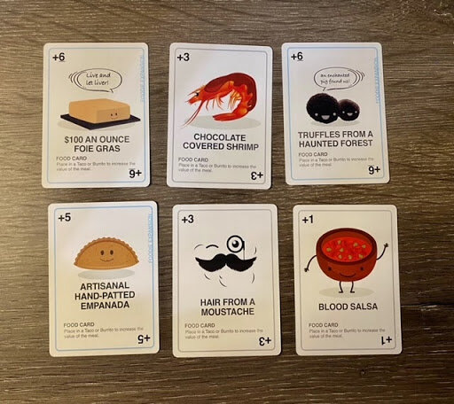 Selection of Taco vs Burrito food ingredient cards laid out on a wooden table, including humorous items like chocolate-covered shrimp, haunted forest truffles, empanadas, and blood salsa.