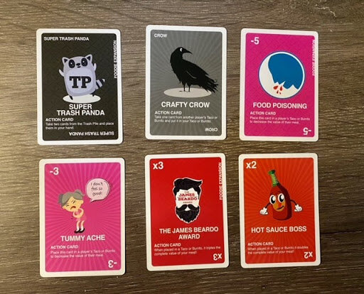 Set of Taco vs Burrito action cards arranged on a wooden surface, featuring playful illustrations and effects such as Super Trash Panda, Crafty Crow, Food Poisoning, and Hot Sauce Boss.
