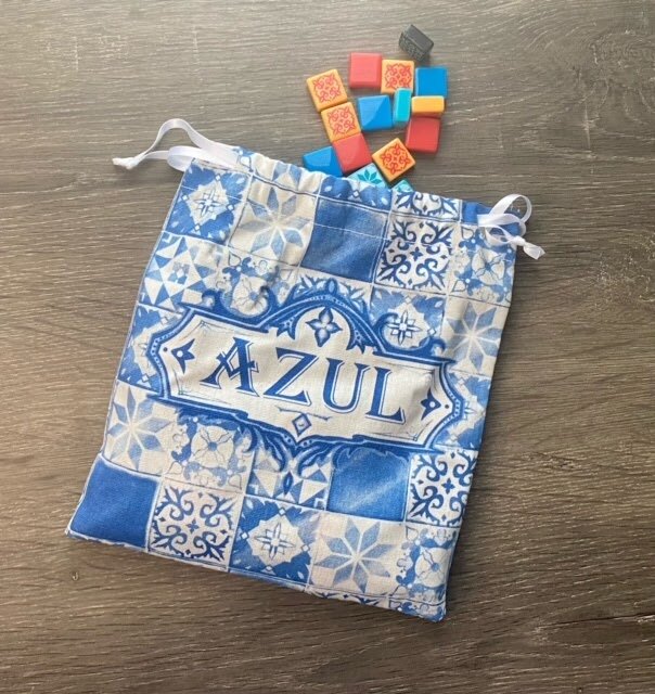 Cloth draw bag with a blue and white tile pattern and the Azul logo, shown with several colorful resin tiles spilled out on a wooden table.