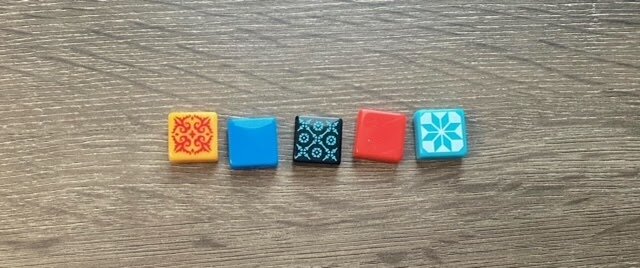 Close-up of five Azul resin tiles in different colors and patterns, arranged in a row on a wooden table.