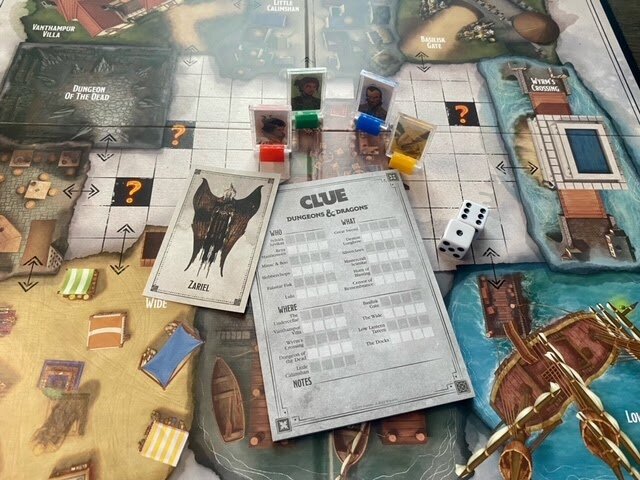 Top-down view of the Clue: Dungeons & Dragons game board fully set up, showing character standees, dice, suspect cards, and the adventure-themed map layout.