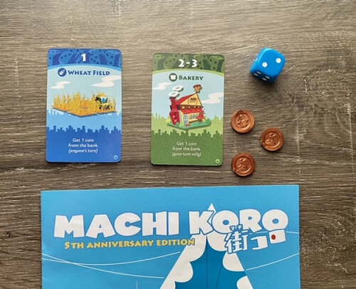 Starter cards from Machi Koro laid out on a table, including Wheat Field and Bakery cards, alongside a blue die and several game coins.
