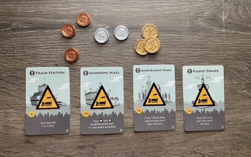 Landmark cards from Machi Koro, including Train Station, Shopping Mall, Amusement Park, and Radio Tower, displayed with game coins above them.