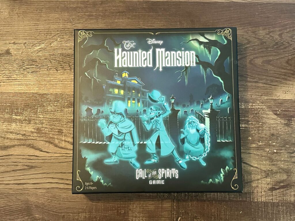 The game box for Haunted Mansion Call of the Spirits Game pictures a mansion in the background, graveyard in the middle ground and 3 cartoon style hitchhiking ghosts in the foreground. 