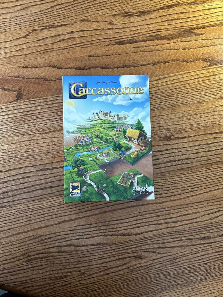 A board game box reads Carcassonne on the top. Tiles show a green village being put together in the foreground and a castle in the background. Z-Man Games logo is pictured on the front right of the box. 