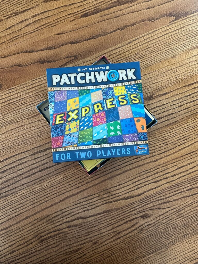 The board game Patchwork Express sits on a wooden table. The box top is off center, revealing some of the components below. The box reads Patchwork Express for Two Players on it. The icon for Lookout Games is in the bottom right corner. 