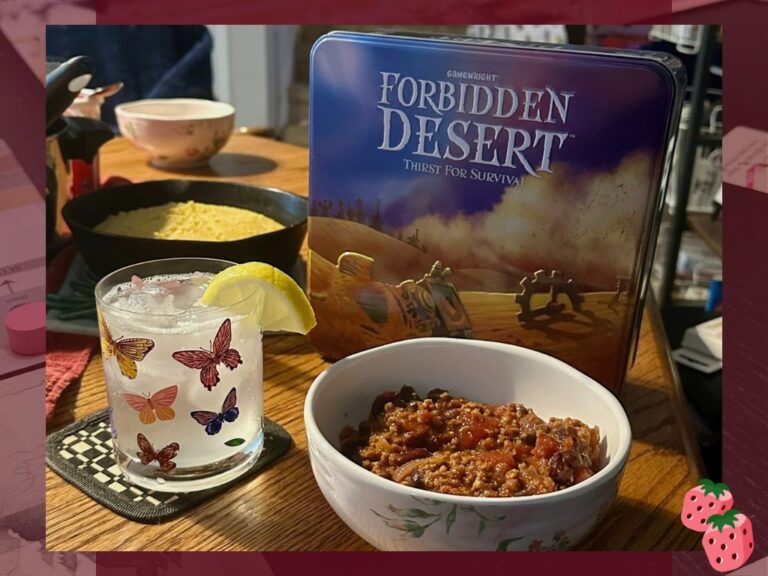 A close up shot of cornbread and chili in a bowl with a clear drink behind it. The game Forbidden Desert is in the background.