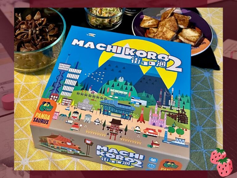 A Machi Koro 2 game box is on display on top of a yellow table cloth. Behind the box is an array of Japanese food.