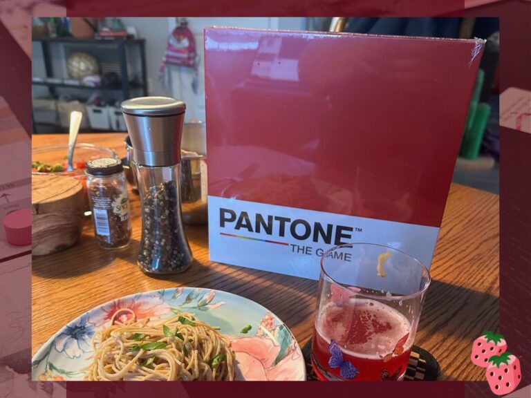On a wooden table stands the game Pantone the Game, next to a pepper mill. In front is a floral plate with spaghetti on it, topped with green leaves. Next to the plate is a glass covered in butterflies with a red drink inside.
