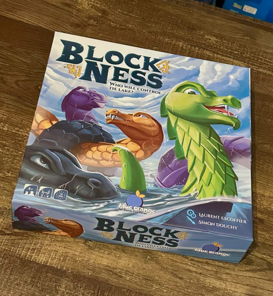 Block Ness board game box on a wooden table, featuring colorful illustrated lake monsters emerging from the water. The box shows the title “Block Ness” with the tagline “Who will control the lake?” and indicates it is for ages 8+, 2–4 players, with a 15-minute playtime.