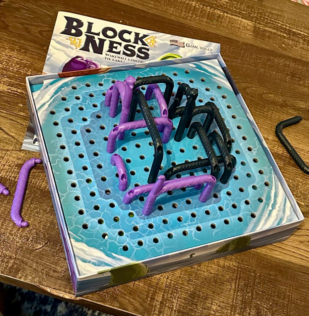 Opened Block Ness board game box on a wooden table, showing the perforated lake board insert with numerous holes and several curved plastic monster neck pieces in purple and black placed on top. The rulebook is partially visible beneath the insert.