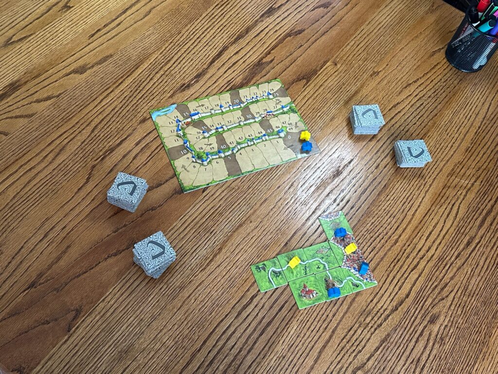 A Carcassonne game in progress on a wooden table. The score board is on the top of the frame, showing two meeples on the 0 starting space. Upside down tiles lay to the left and the right. In the bottom center show tiles, green side up, piecing together a river and a village. 