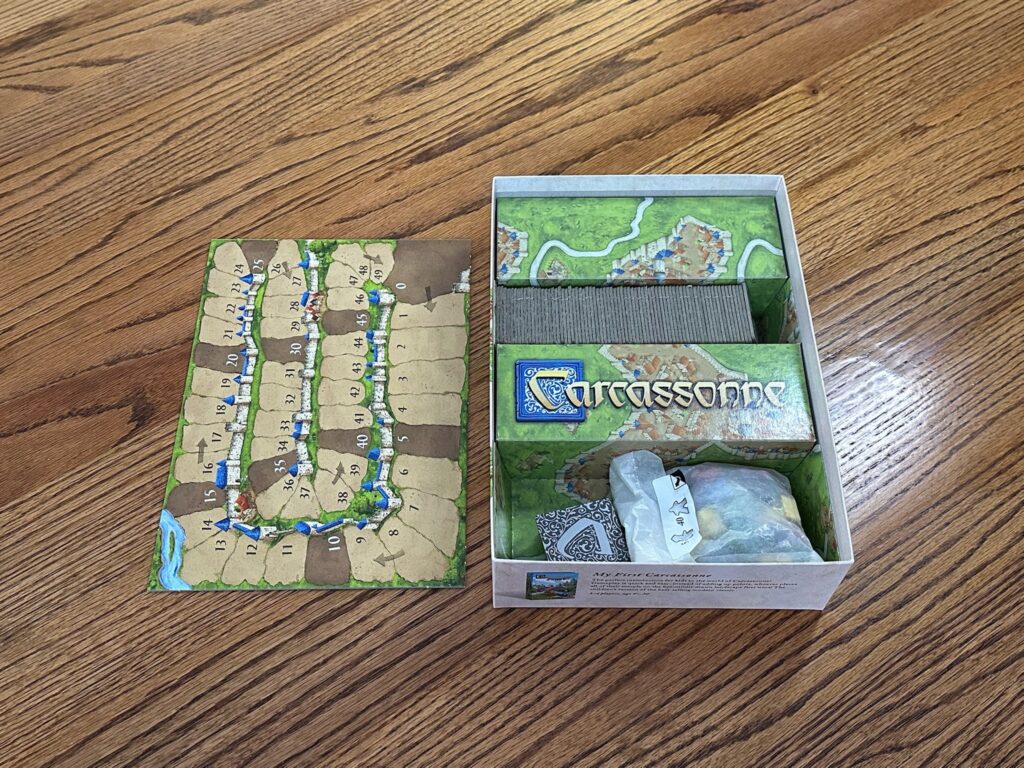 An open Carcassonne game box sits on a wooden surface. A game board is on the left hand side with numbers 0-49. In the box you can see piles and meeples, ready to be played. 
