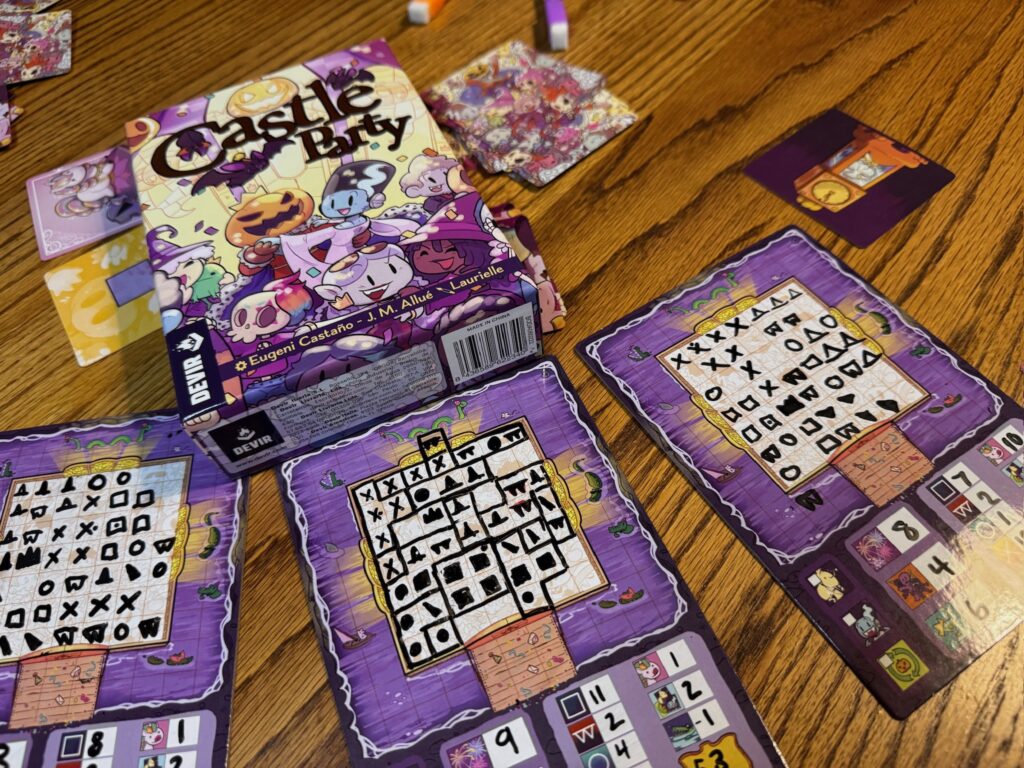 A wooden tabletop holds the game box that reads Castle Party. in front of the game box and 3 roll and write boards with boxes and symbols on the, to the right is a picture of a grandfather clock. 
