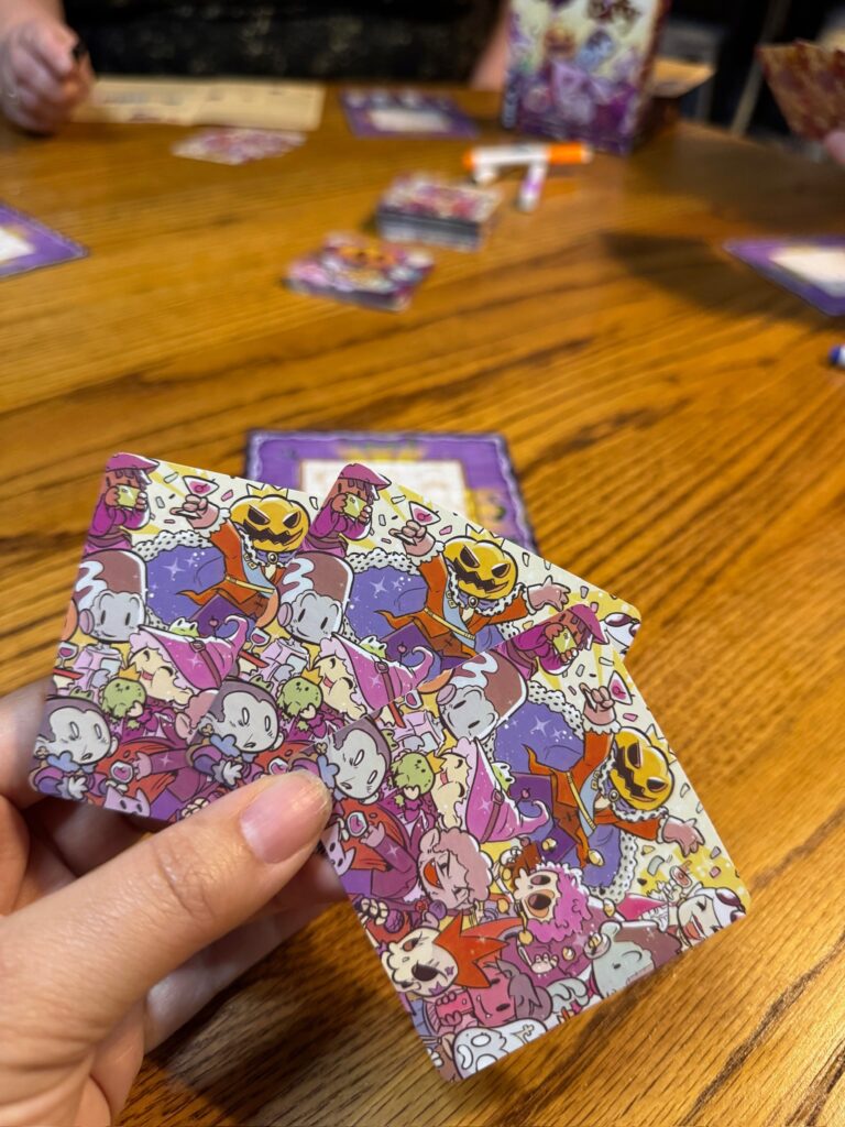 A hand holds 3 cards with Halloween characters pictured on them. In the background you can see others playing the game Castle Party. 