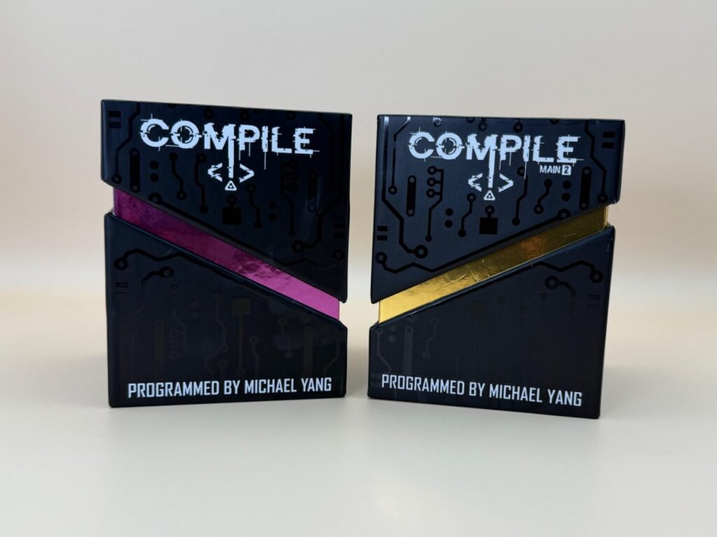 Two boxes for Compile 1 & 2 sit side by side. The boxes are matte black with embossed motherboard-esque designs. Each box has an angled top with a foil decoration underneath. "Programmed by Michael Yang" is scrolled across the bottom.