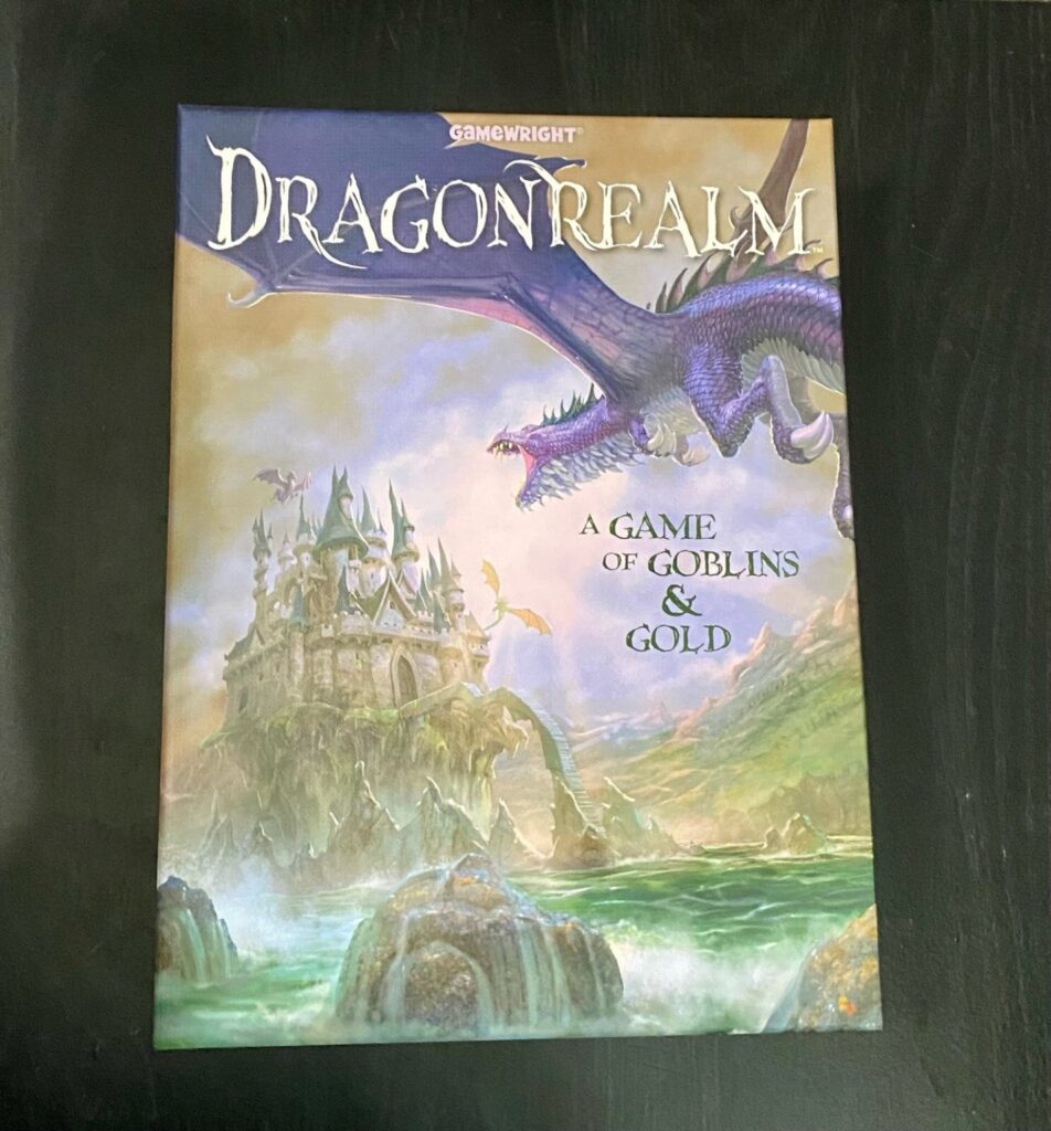 A game box with the words DragonRealm written at the top. A purple dragon flies towards a castle above the sea. The works A Game of Goblins and Gold are written in the right center of the box top.