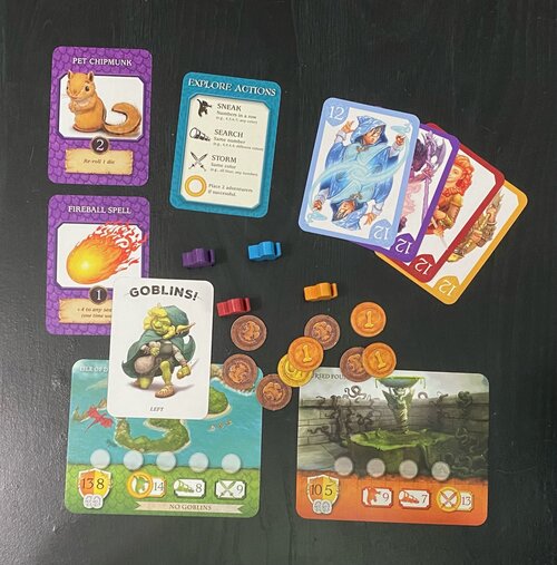 The items of the game are spread out on a table. Items include numbered cards, maples, coins, location cards, a pet chipmunk card, a fireball spell card and a goblins card.