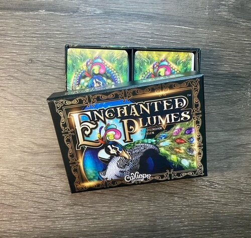 A box reads Enchanted Plumes on the top center, picturing a peacock in the center. Box top is partially opened, revealing 2 decks or cards inside.