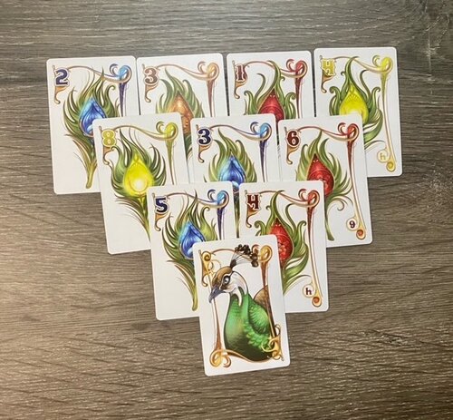 A full layout of 10 Enchanted Plumes cards are shown in an inverted pyramid. The top card features a peacock's body. The additional 9 cards feature feathers of different colors and numerical values.