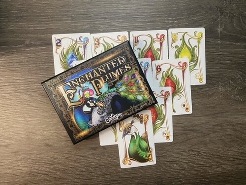 The Enchanted Plumes game box is pictured on top of an array of cards from the game.