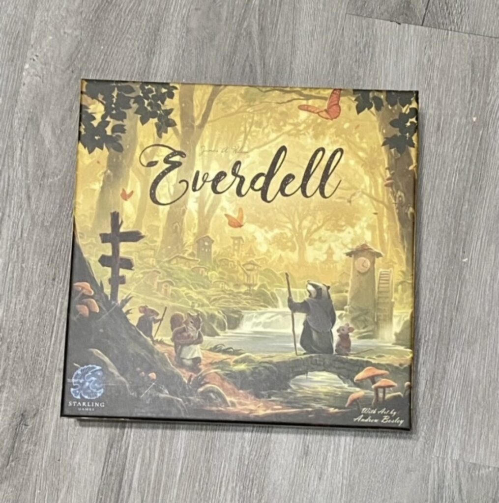 A game box pictured a scene in the woods has the word Everdell written at the top center. Also on the box is the Starling Games logo in the bottom left. A badger standing upright and a mouse are on the right hand side of the picture, looking at a small waterfall. 