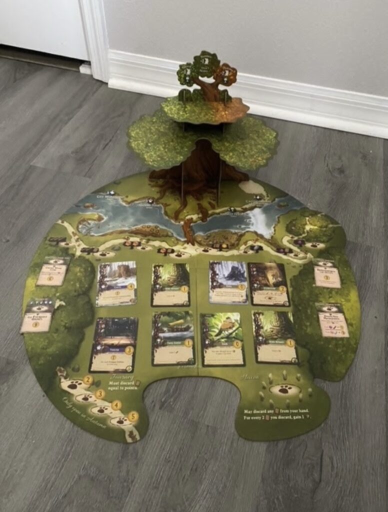 A game board with a woodlands theme depicts a river below a 3D, 3 level tree. Game cards are placed around the board. 