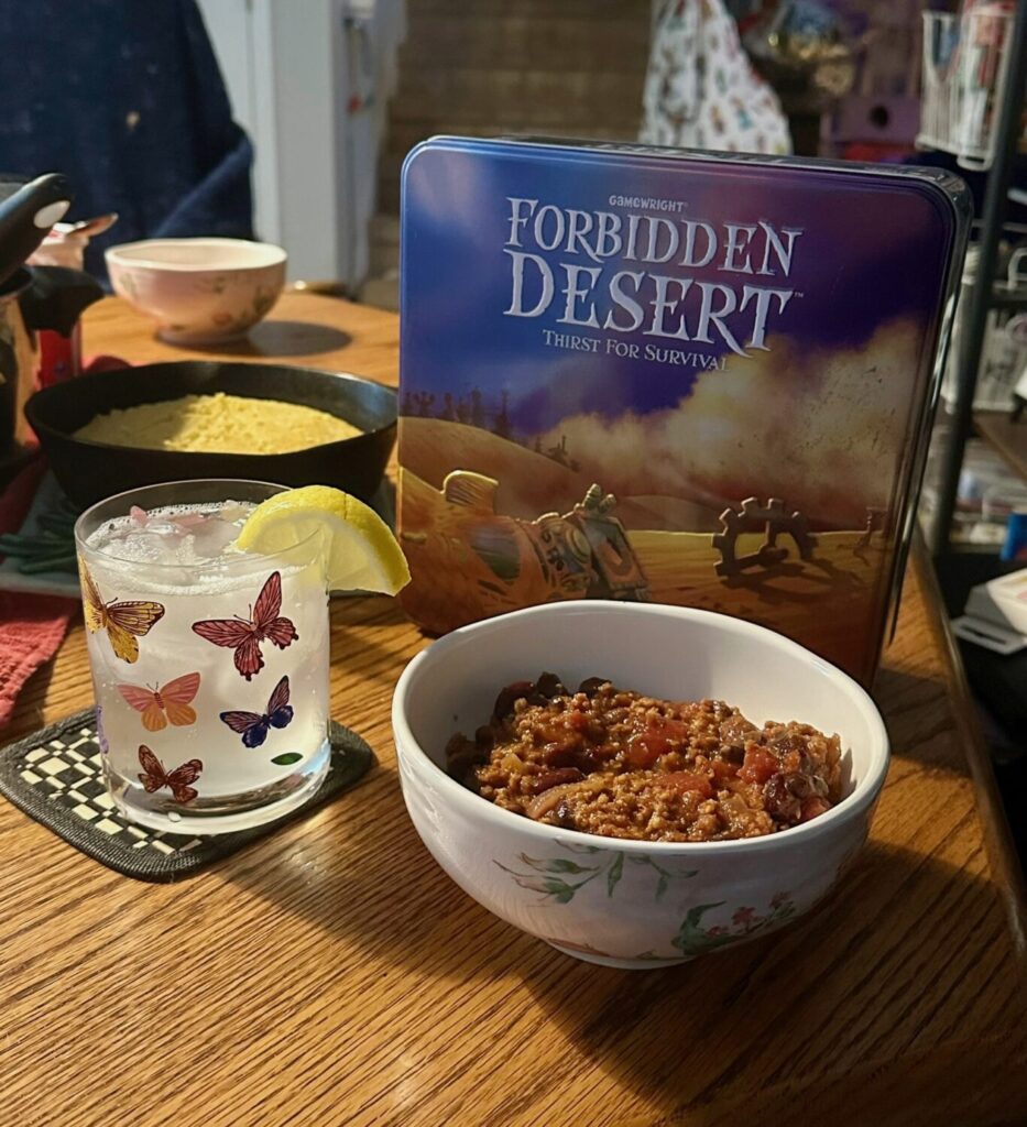 A close up shot of cornbread and chili in a bowl with a clear drink behind it. The game Forbidden Desert is in the background.