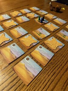 Cardboard desert tiles with desert images on them are set on a wooden surface. Several of the tiles are covered with a picture of a pile of sand over them.
