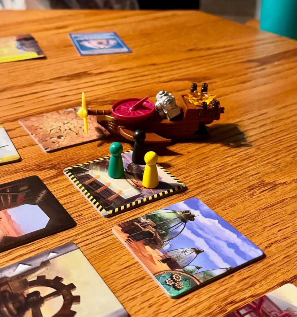 Components for Forbidden Desert are featured, include gameplay tiles, 3 meeples of different colors, and various plastic pieces making up a machine are in the background.